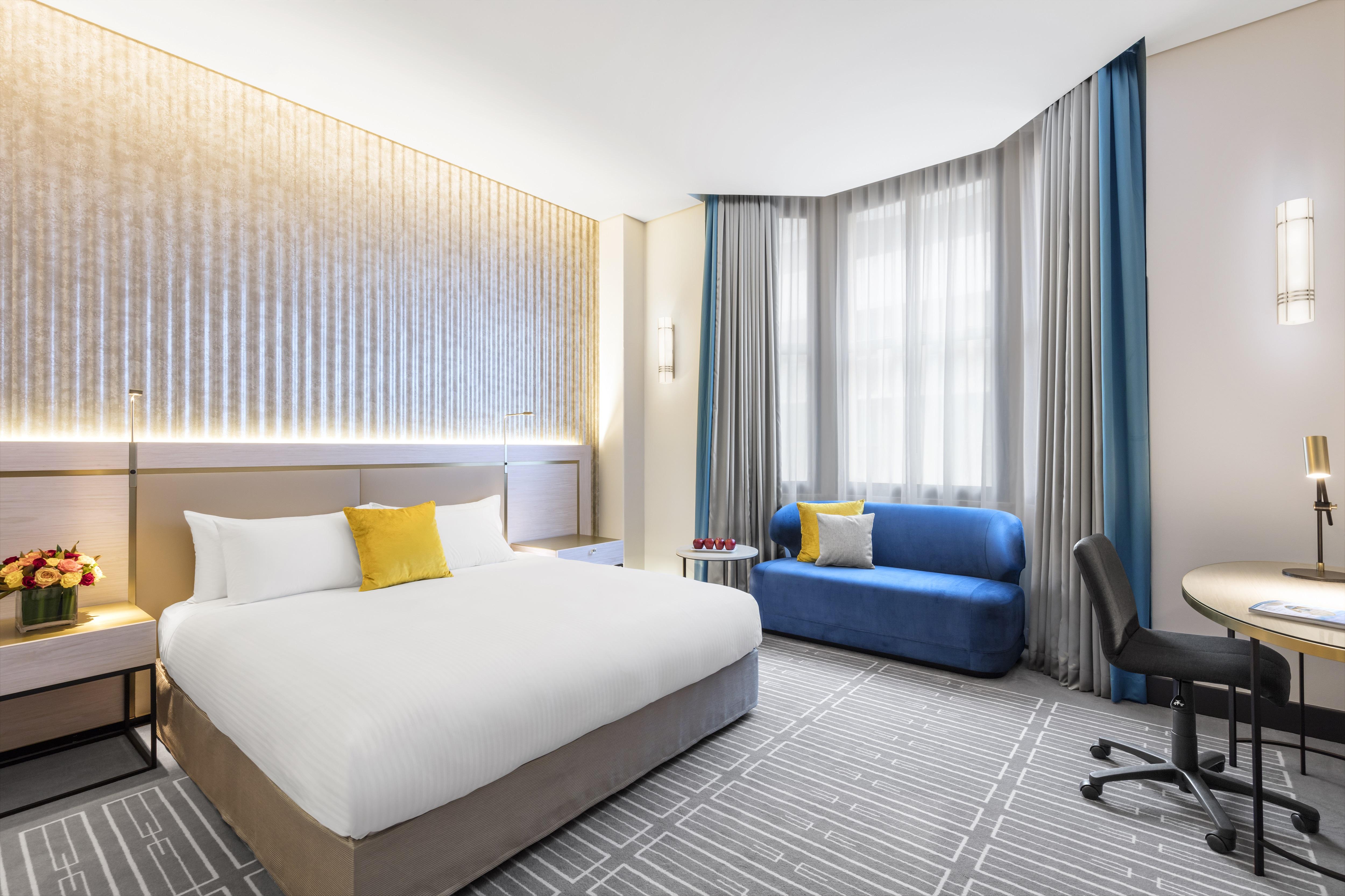 Radisson Blu Plaza Hotel Σίδνεϊ Εξωτερικό φωτογραφία The photo shows a modern hotel room featuring a large bed with white linens and a yellow accent pillow. To the side, there is a blue sofa and a small round table with a chair positioned next to it. The room is well-lit, with natural light coming thro