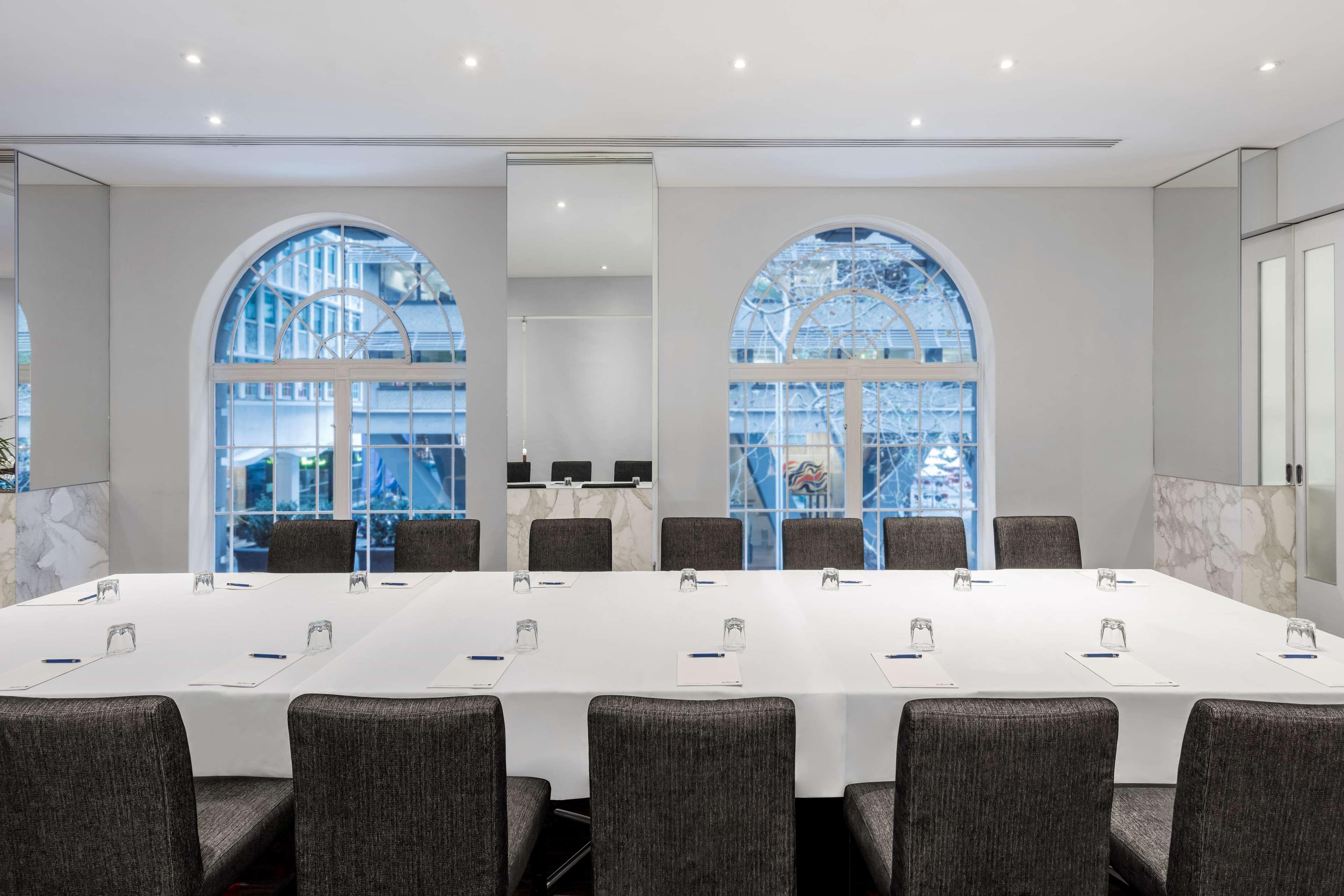 Radisson Blu Plaza Hotel Σίδνεϊ Εξωτερικό φωτογραφία The image shows a conference room set up for a meeting. It features a long table covered with a white tablecloth, arranged with several place settings and water glasses. Around the table are black, padded chairs. The room has large arched windows tha