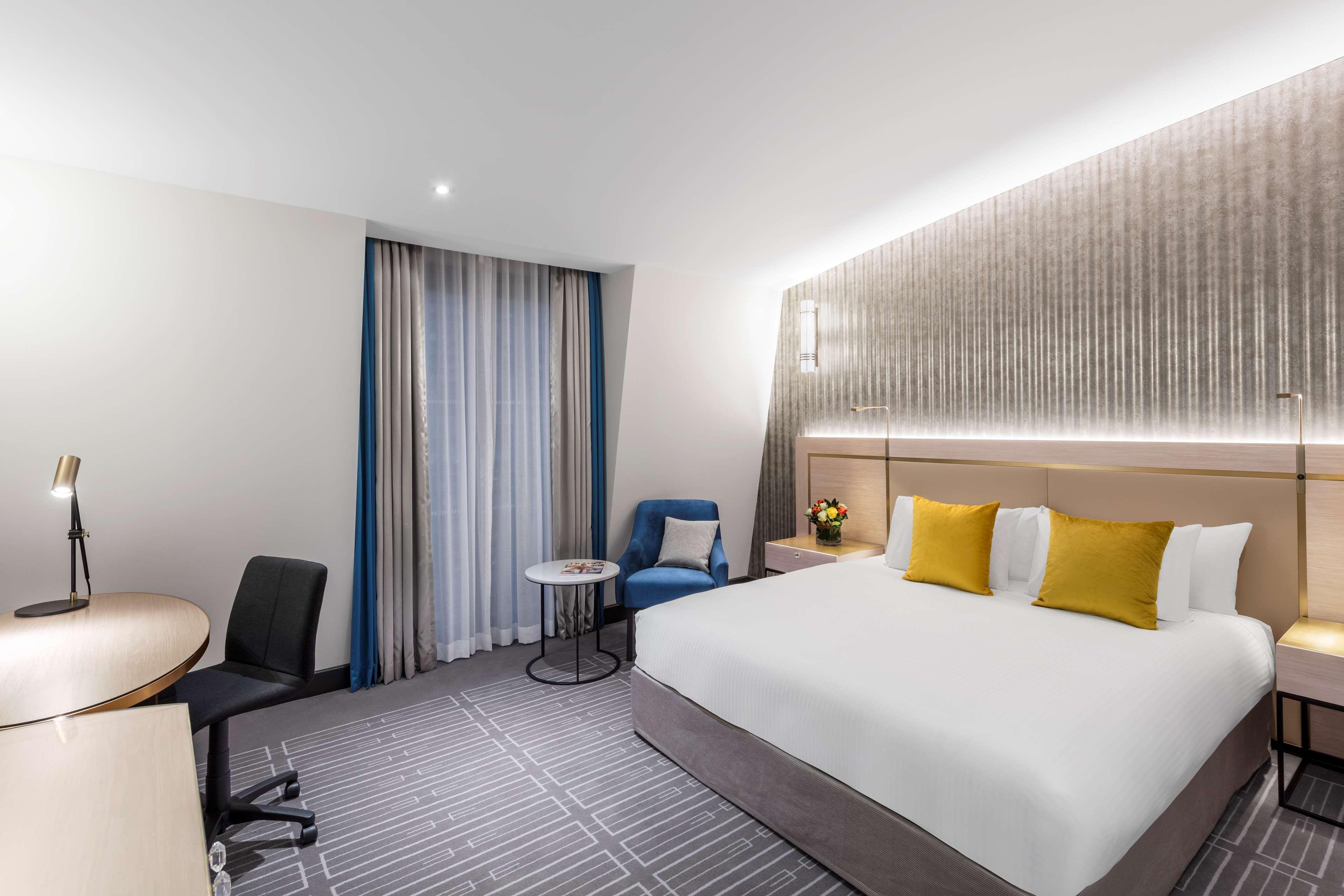 Radisson Blu Plaza Hotel Σίδνεϊ Εξωτερικό φωτογραφία The photo shows a modern hotel room featuring a large bed with white linens and yellow accent pillows. There's a plush blue chair next to a small round table. To one side, a desk with a black office chair is positioned, likely for work or study. The 