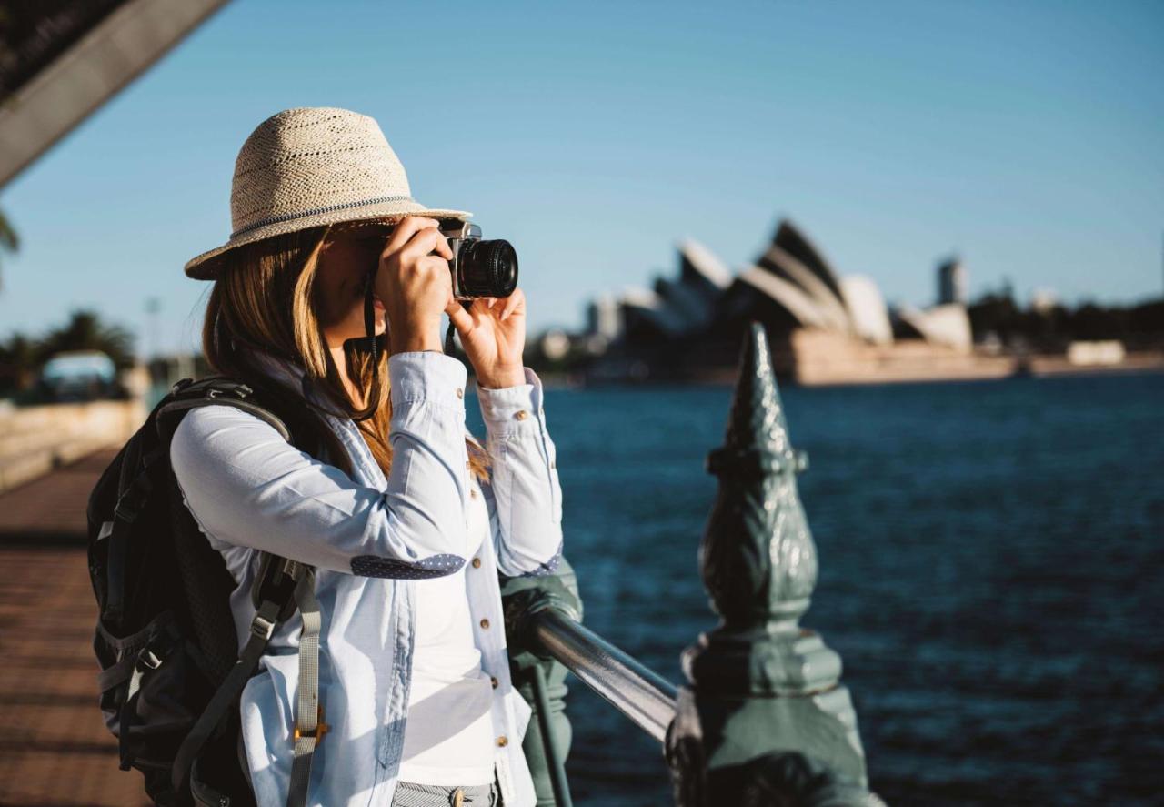 Radisson Blu Plaza Hotel Σίδνεϊ Εξωτερικό φωτογραφία The photo shows a woman standing by a railing near a body of water, taking a picture with a camera. She is wearing a light-colored hat and a backpack, dressed in a casual outfit. In the background, the iconic Sydney Opera House is visible, along with