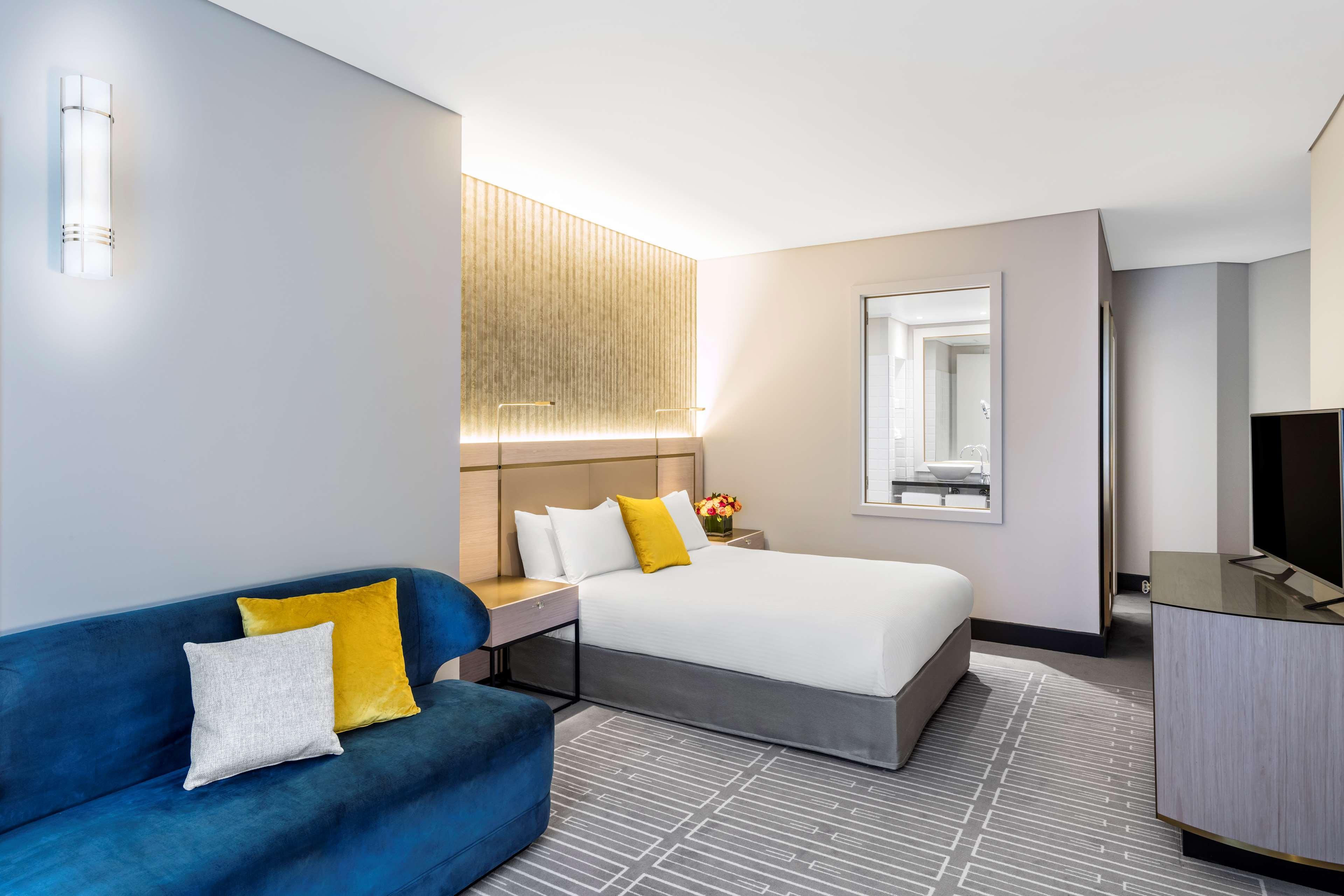 Radisson Blu Plaza Hotel Σίδνεϊ Εξωτερικό φωτογραφία The photo shows a modern hotel room. It features a double bed with white bedding and a grey upholstered base. To the left, there is a blue sofa accompanied by decorative pillows in yellow and white. A small wooden side table sits beside the sofa. The