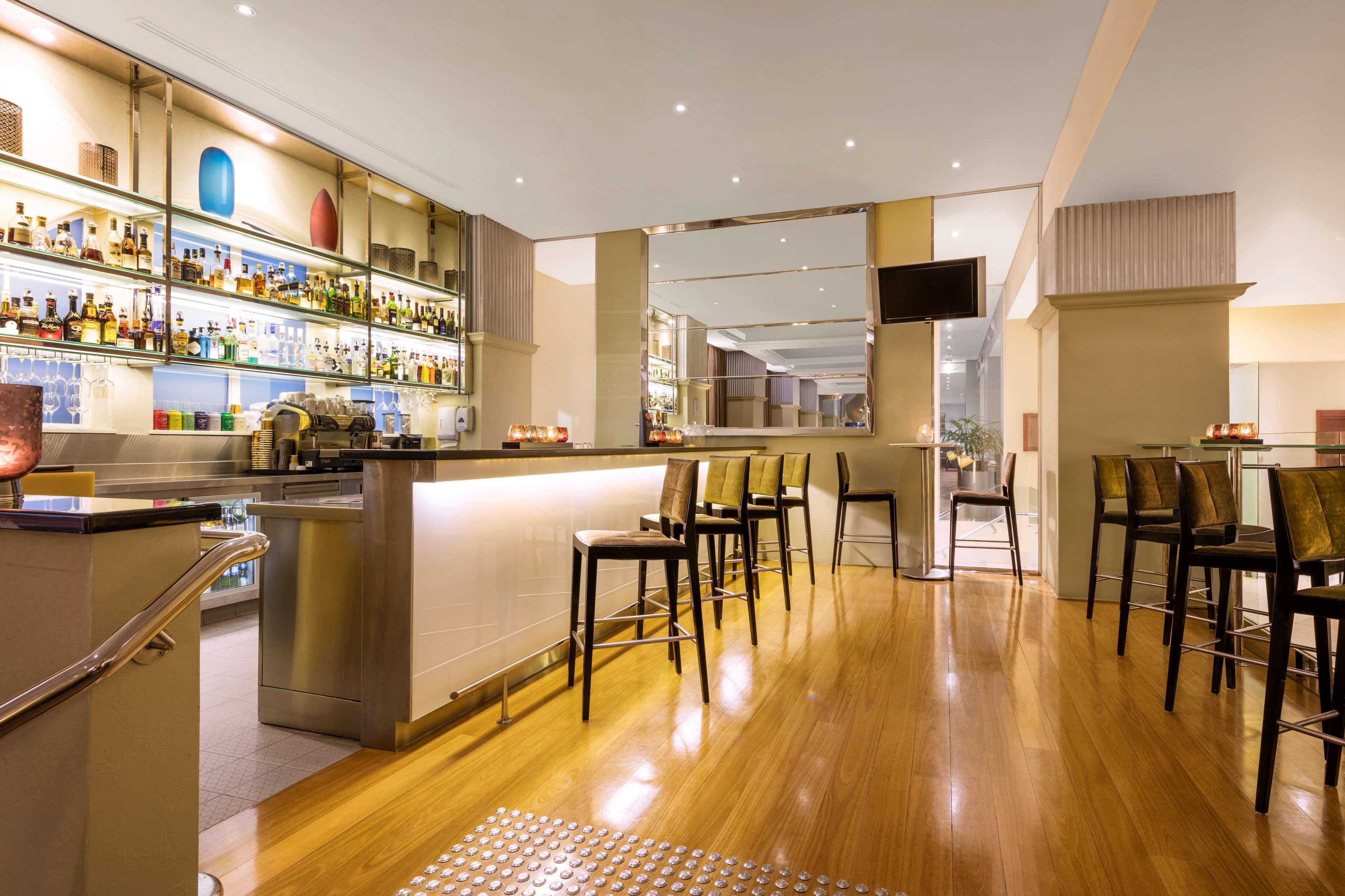 Radisson Blu Plaza Hotel Σίδνεϊ Εξωτερικό φωτογραφία The photo shows the interior of a modern bar or lounge area. There is a sleek bar counter with several high-backed stools arranged around it. Behind the bar, various bottles of liquor and decorative items are displayed on shelves, illuminated by soft