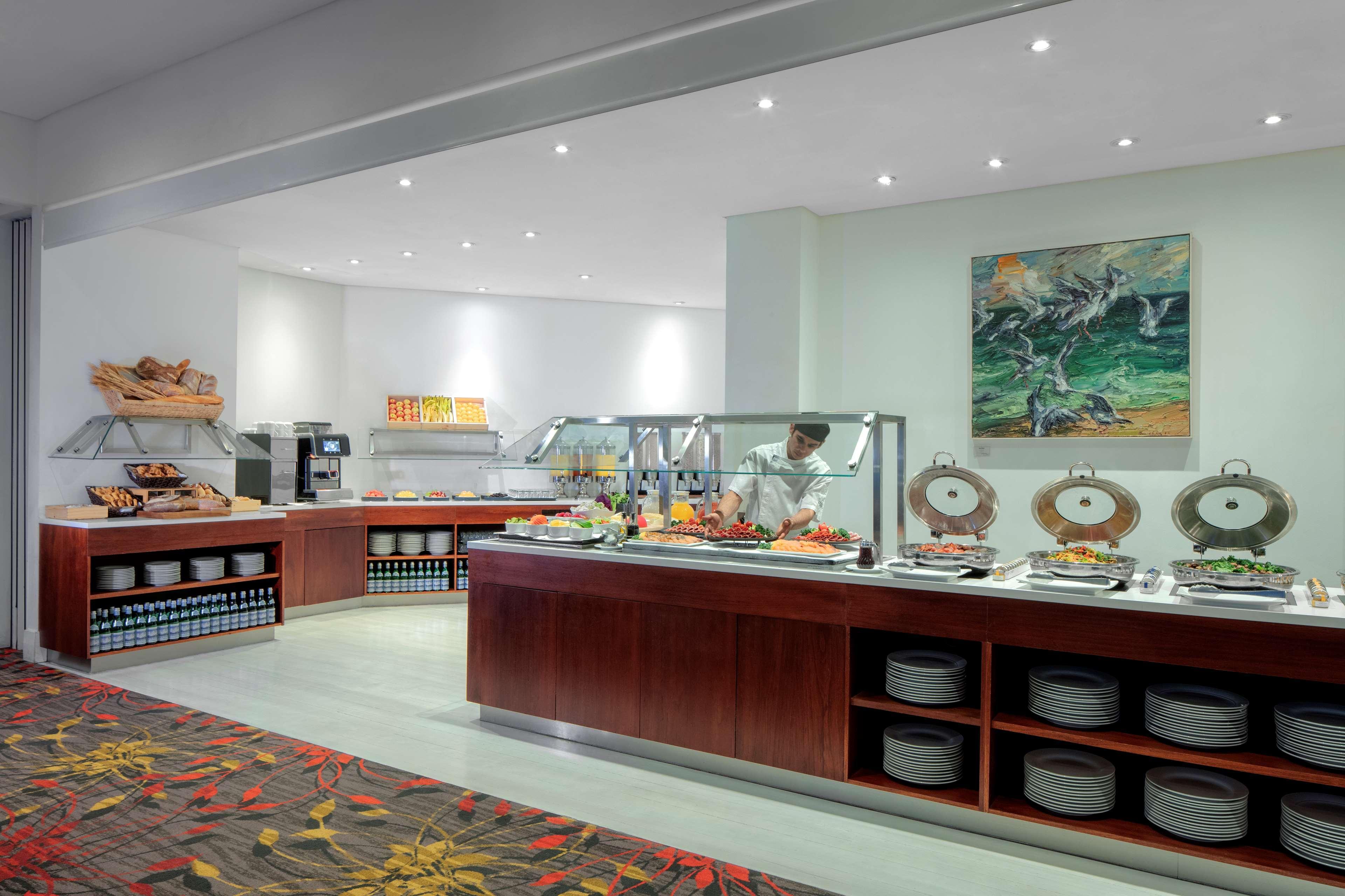 Radisson Blu Plaza Hotel Σίδνεϊ Εξωτερικό φωτογραφία The photo shows a buffet area in a dining space. There are several food stations arranged along a wall, featuring various dishes and offerings. A chef is visible behind the buffet, preparing food. The design includes wooden counters and shelves for s