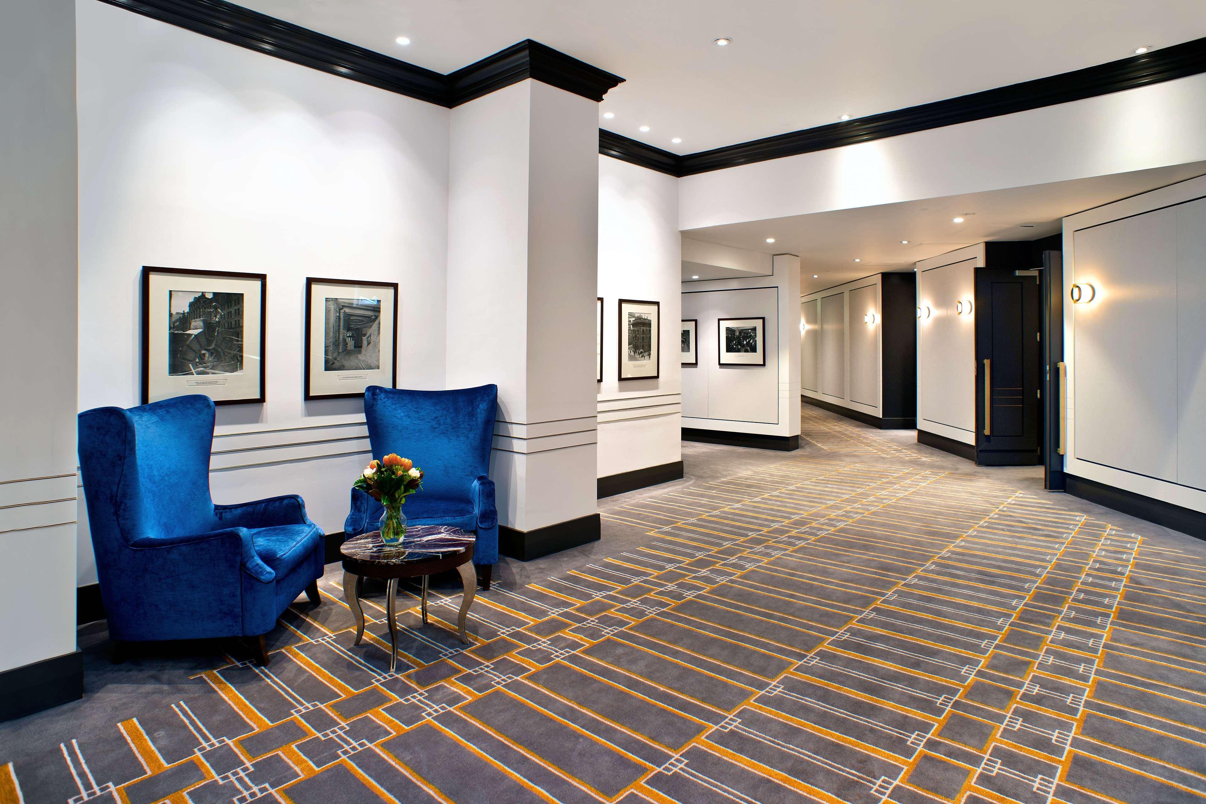 Radisson Blu Plaza Hotel Σίδνεϊ Εξωτερικό φωτογραφία The photo shows a modern hotel or building hallway. On one side, there are two vibrant blue armchairs arranged around a small table with a vase of flowers on it. The walls feature a few framed pictures, possibly black and white photographs. The floor