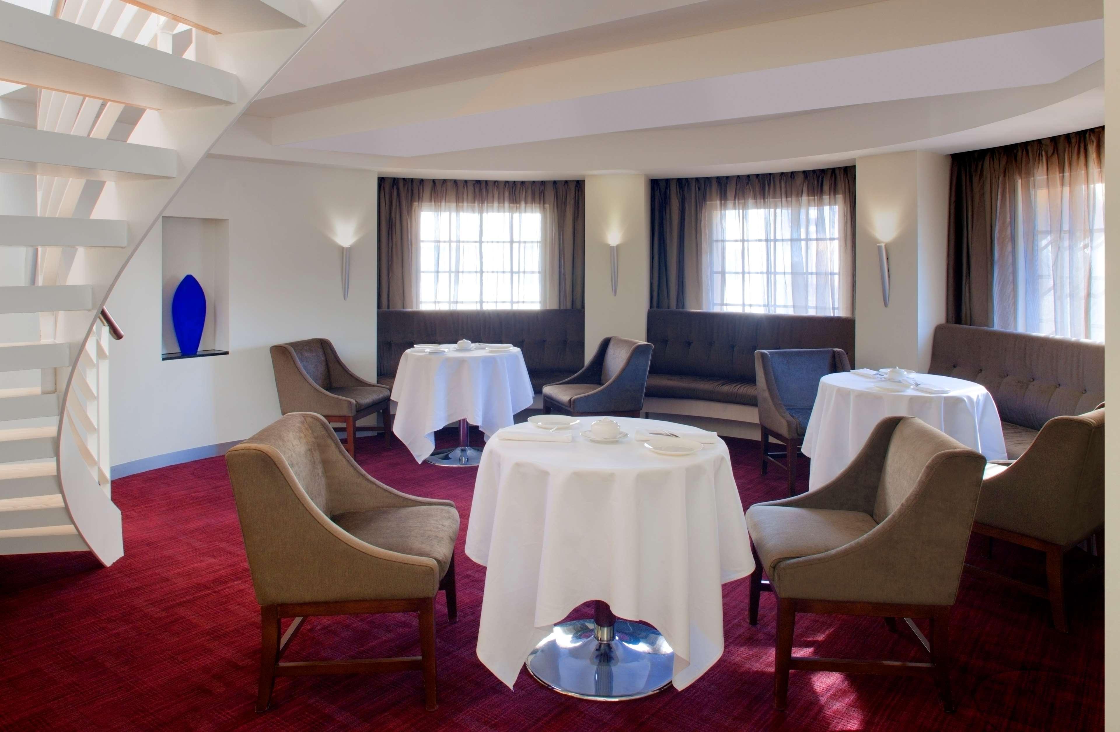 Radisson Blu Plaza Hotel Σίδνεϊ Εστιατόριο φωτογραφία The photo shows a stylish dining area with a modern design. It features several round tables, each draped with white tablecloths. The seating consists of comfortable chairs with a neutral color, complemented by a plush bench along one wall. The space