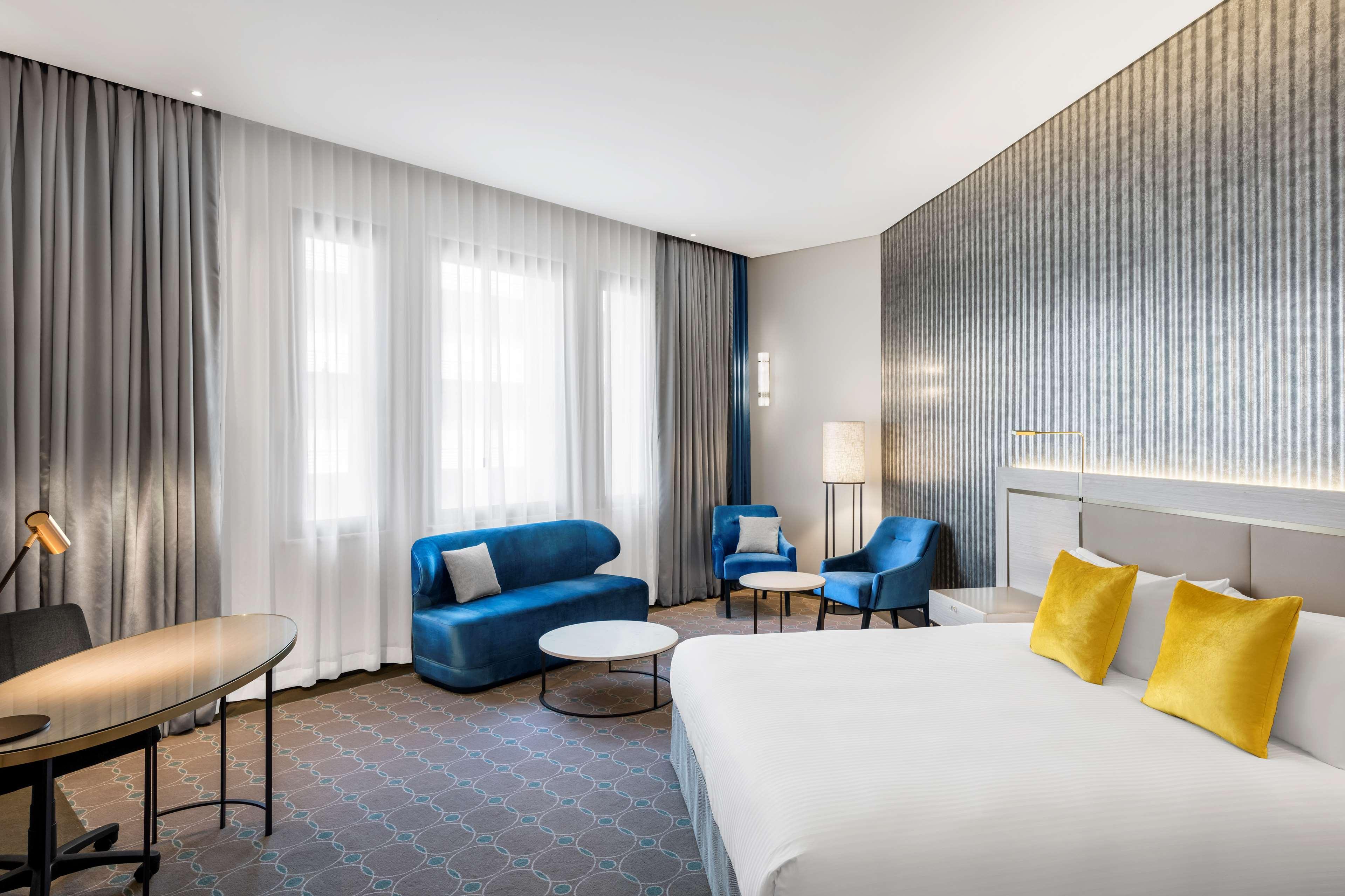 Radisson Blu Plaza Hotel Σίδνεϊ Εξωτερικό φωτογραφία The photo depicts a modern hotel room featuring a cozy and stylish design. On one side, there is a large bed with white linens and bright yellow accent pillows. The room includes a small seating area with a blue sofa and two blue chairs, arranged aro
