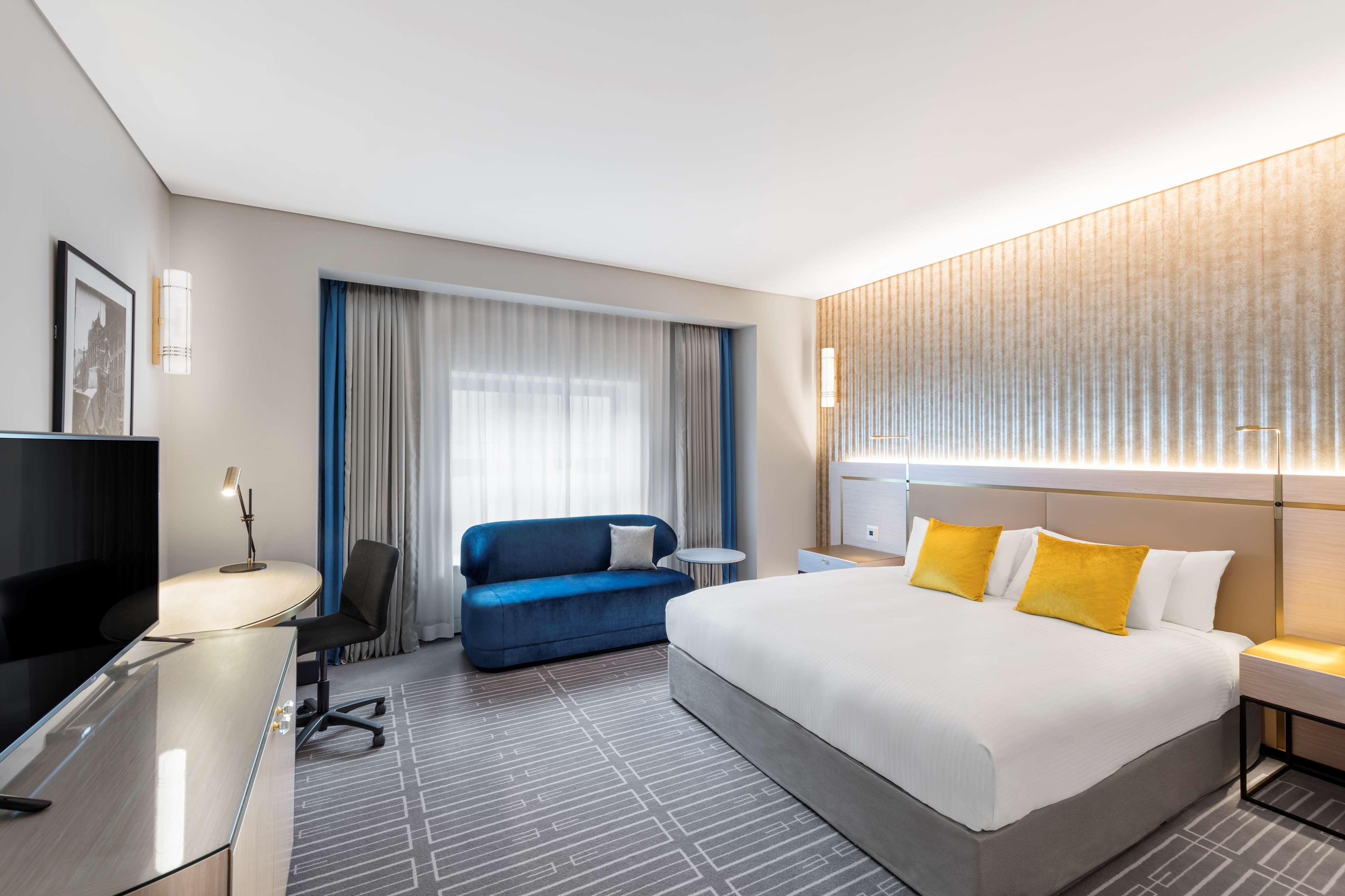 Radisson Blu Plaza Hotel Σίδνεϊ Εξωτερικό φωτογραφία The photo shows a modern hotel room featuring a spacious layout. It has a large bed with white linens and yellow accent pillows. Beside the bed, there is a bedside table and a contemporary lamp. There is a blue sofa and a coffee table near a set of l