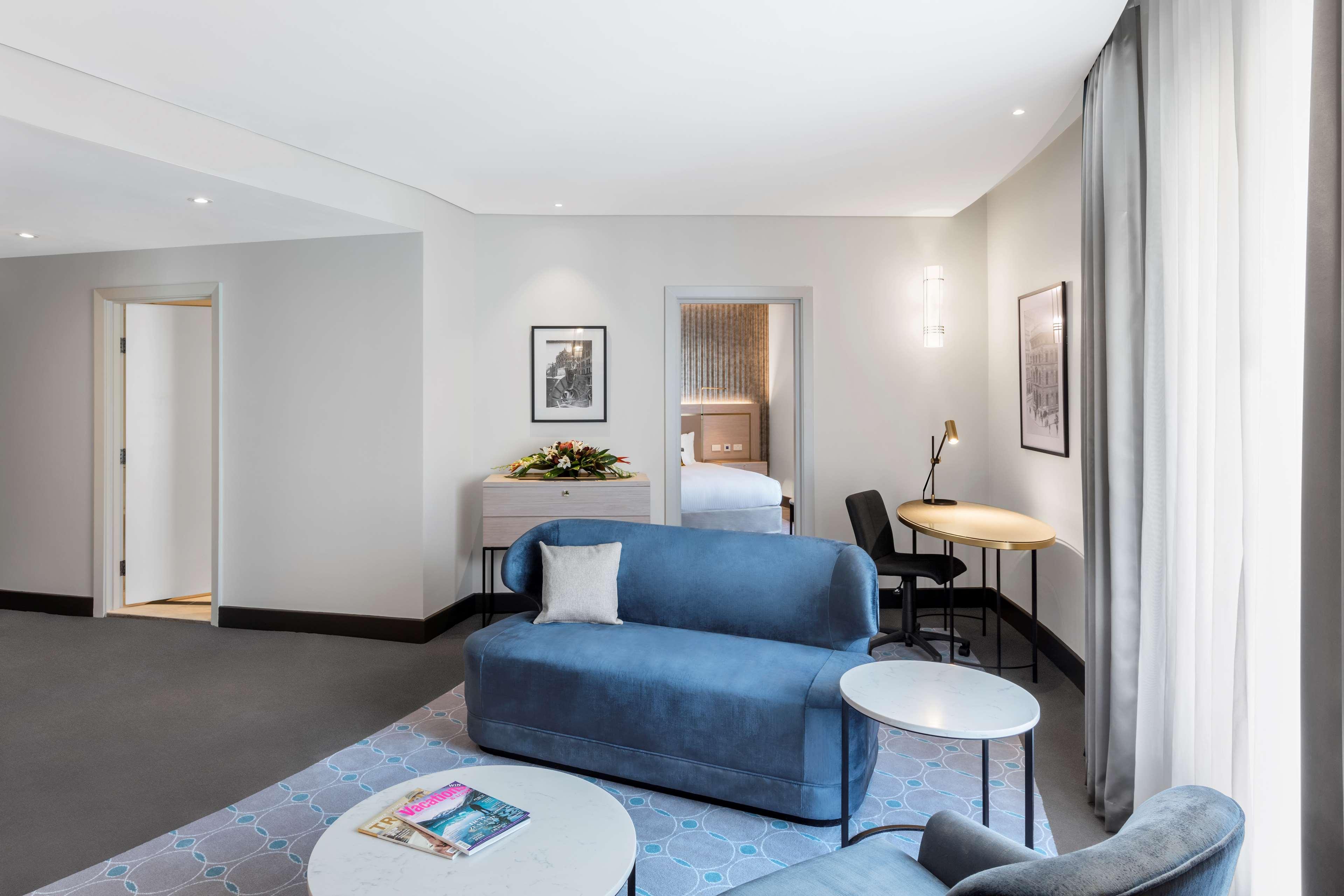 Radisson Blu Plaza Hotel Σίδνεϊ Εξωτερικό φωτογραφία The photo depicts a modern hotel room or suite that features a comfortable and stylish living area. The room includes a light blue sofa complemented by a small round coffee table. There is a decorative pillow on the sofa. To the side, there is a circ