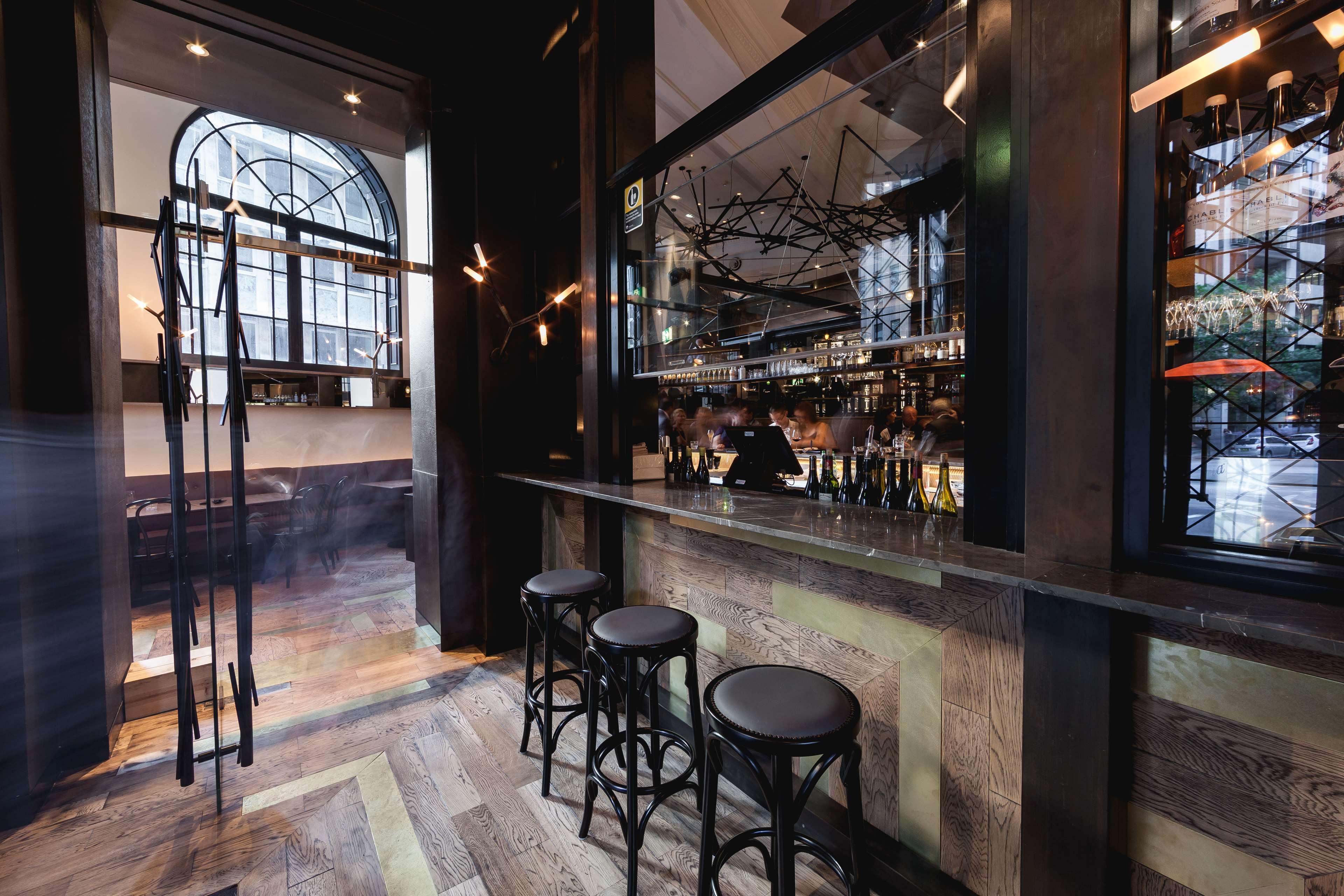 Radisson Blu Plaza Hotel Σίδνεϊ Εξωτερικό φωτογραφία The photo shows the interior of a stylish bar or lounge. There are several tall, black stools arranged in front of a polished bar counter. The bar has a glass backing that showcases a variety of bottles, indicating a well-stocked bar area. The floori