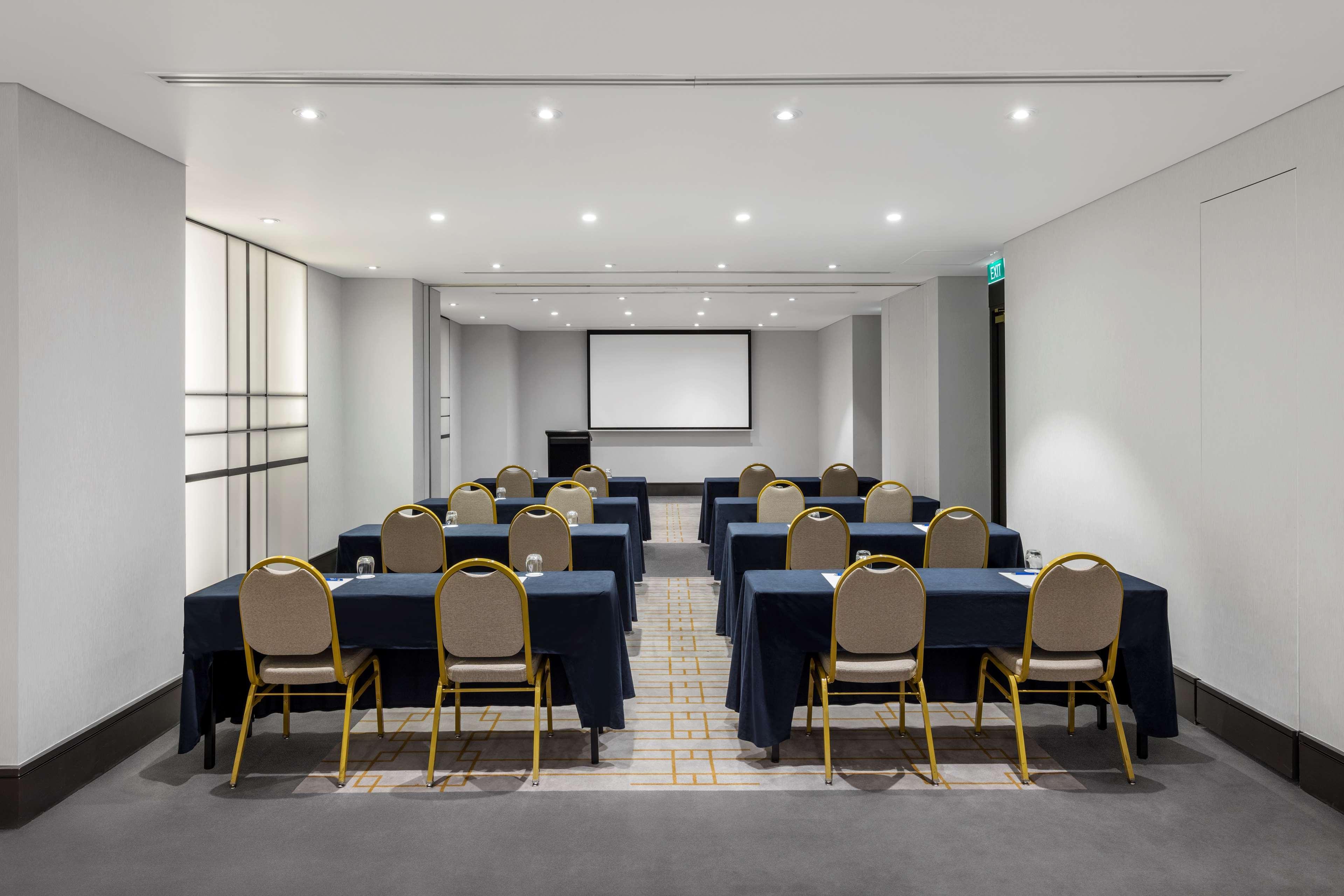 Radisson Blu Plaza Hotel Σίδνεϊ Εξωτερικό φωτογραφία The photo shows a conference room set up for a meeting or presentation. There are several tables arranged in a U-shape, each covered with dark blue tablecloths. Each table has a gold-colored chair positioned at it. At the front of the room, there is 