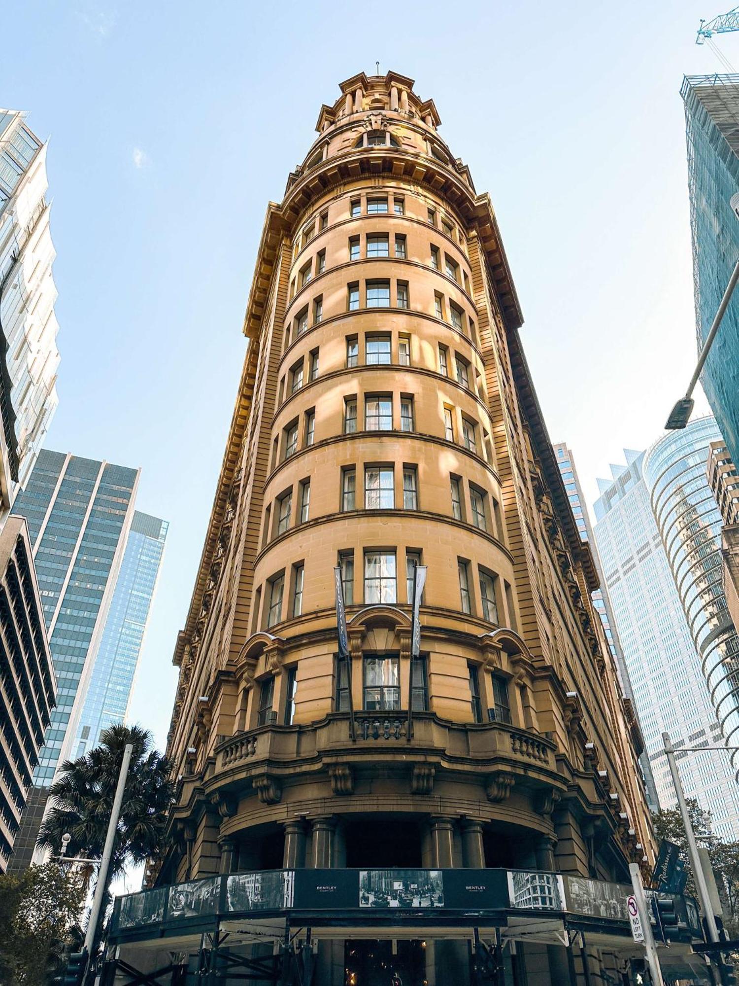 Radisson Blu Plaza Hotel Σίδνεϊ Εξωτερικό φωτογραφία The photo features a tall, historic building characterized by its rounded, cylindrical shape and numerous windows. The facade is ornate with a warm, yellowish-brown color, and the architecture reflects styles from an earlier era. Flanking the buildin