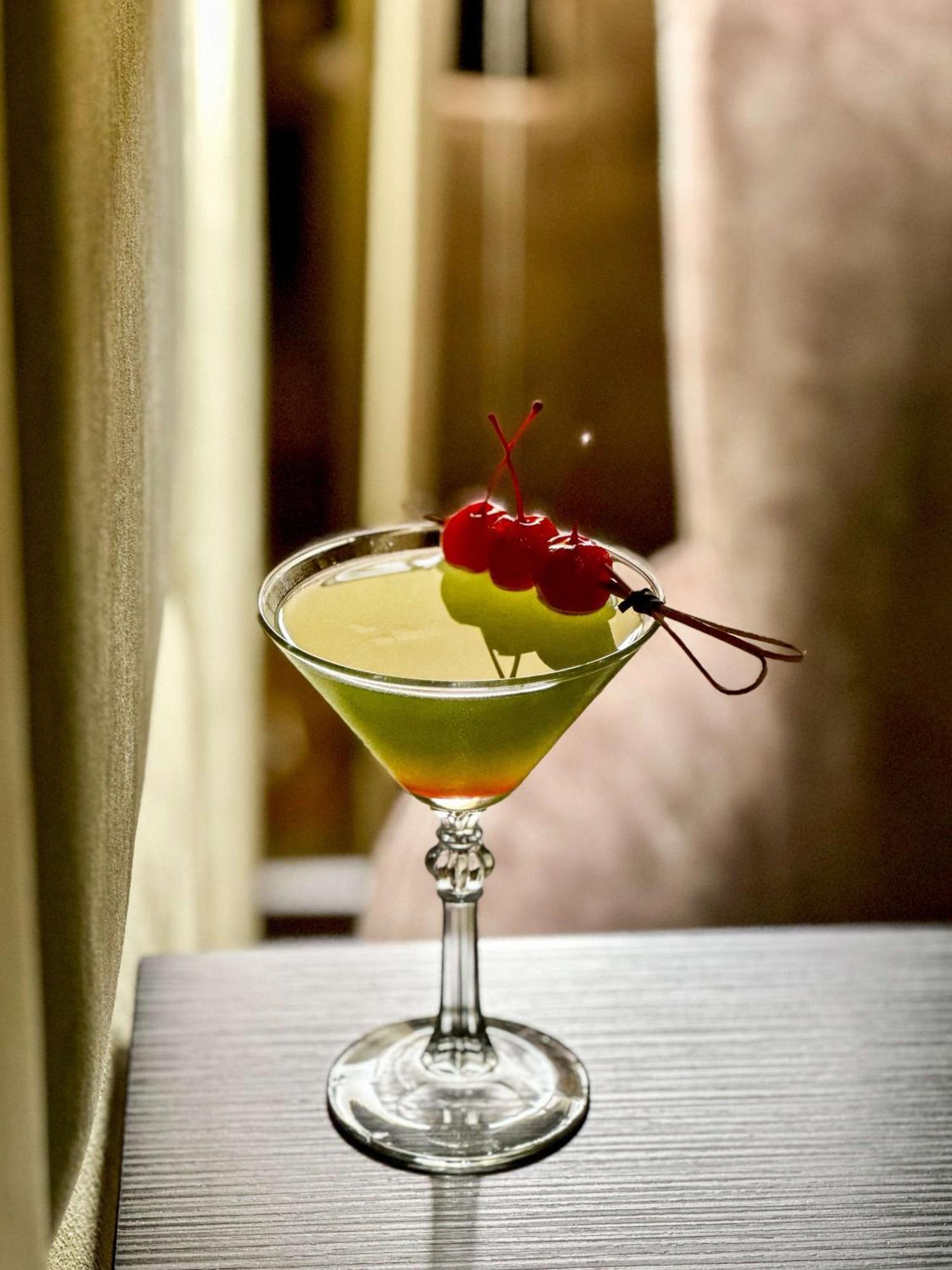 Radisson Blu Plaza Hotel Σίδνεϊ Εξωτερικό φωτογραφία The image shows a cocktail in a classic martini glass. The drink has a vibrant green color and is garnished with two yellow-green fruits (possibly a melon or similar) and three cherries on a skewer. The background appears blurred, creating a soft foc