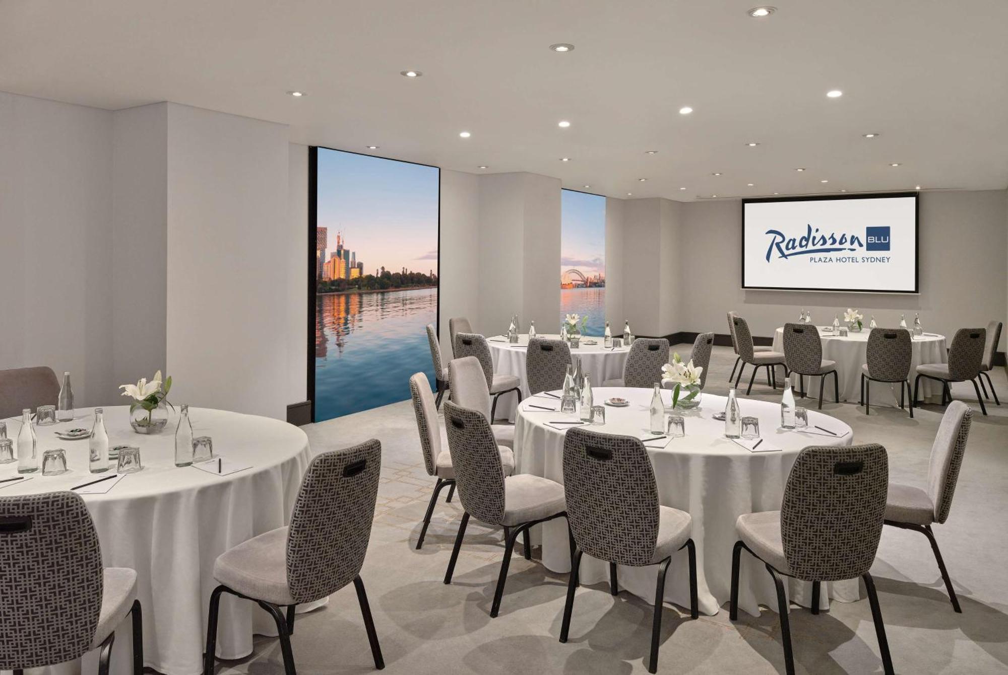Radisson Blu Plaza Hotel Σίδνεϊ Εξωτερικό φωτογραφία The photo shows a modern meeting room at the Radisson Hotel in a city. The room features several round tables set with white tablecloths and gray upholstered chairs. There are small flower arrangements on each table, creating a welcoming atmosphere. 