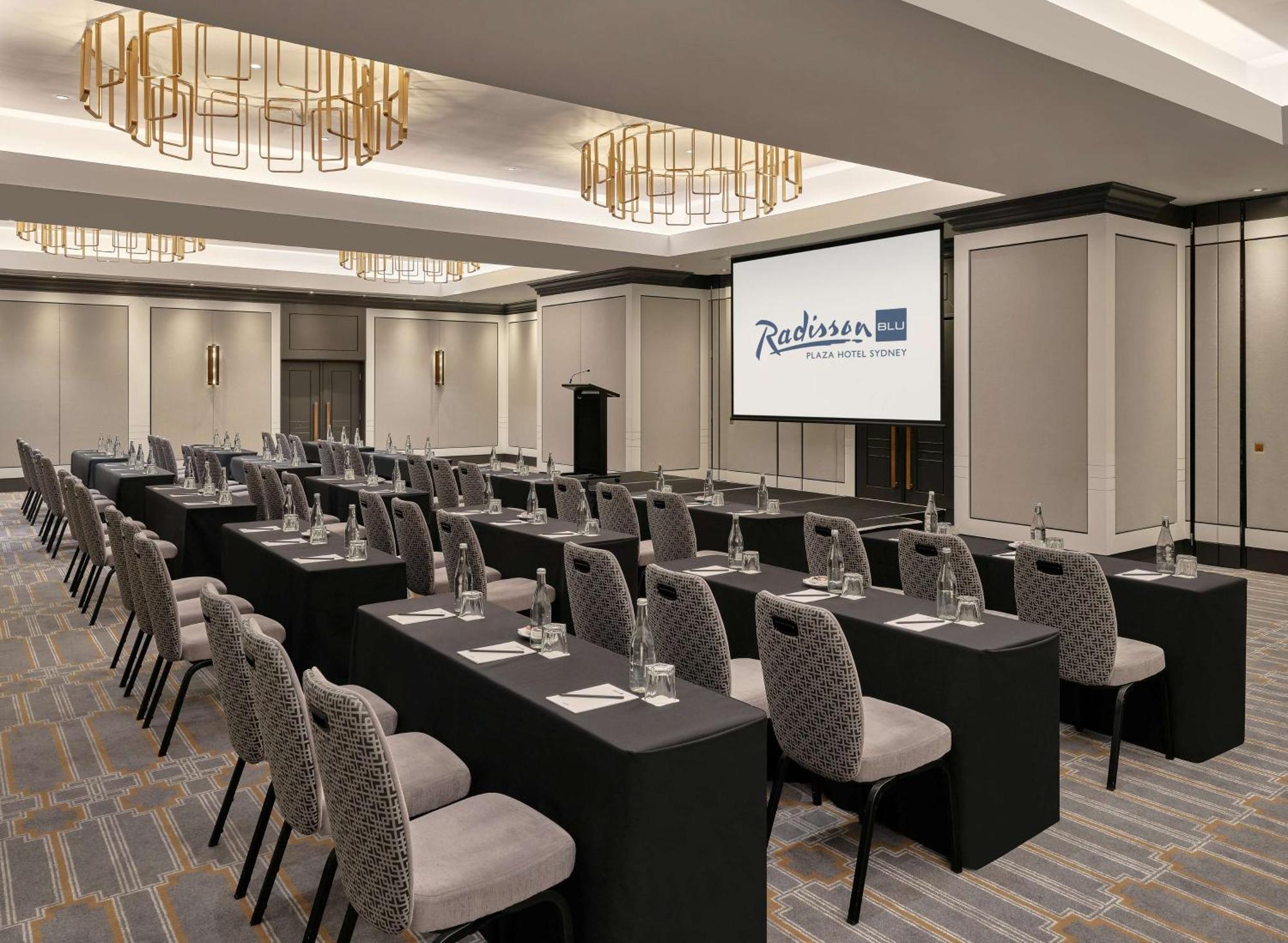 Radisson Blu Plaza Hotel Σίδνεϊ Εξωτερικό φωτογραφία The photo shows a conference or meeting room set up in a hotel, likely a Radisson. The room features several rows of tables covered in black tablecloths, each with a chair beside it. Water glasses are placed on each table, and there appears to be a l